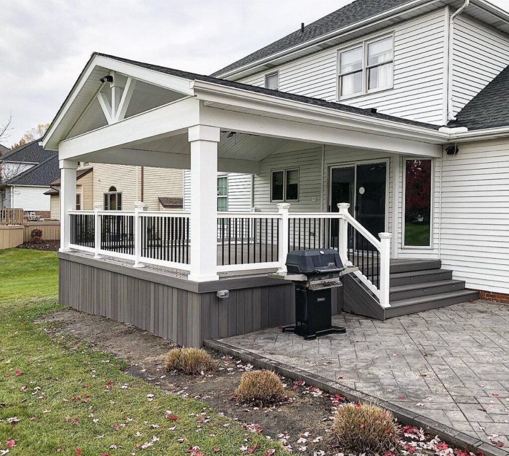 A white covered deck with black railings attached to a white house overlooking a stone patio.