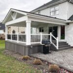 A white covered deck with black railings attached to a white house overlooking a stone patio.