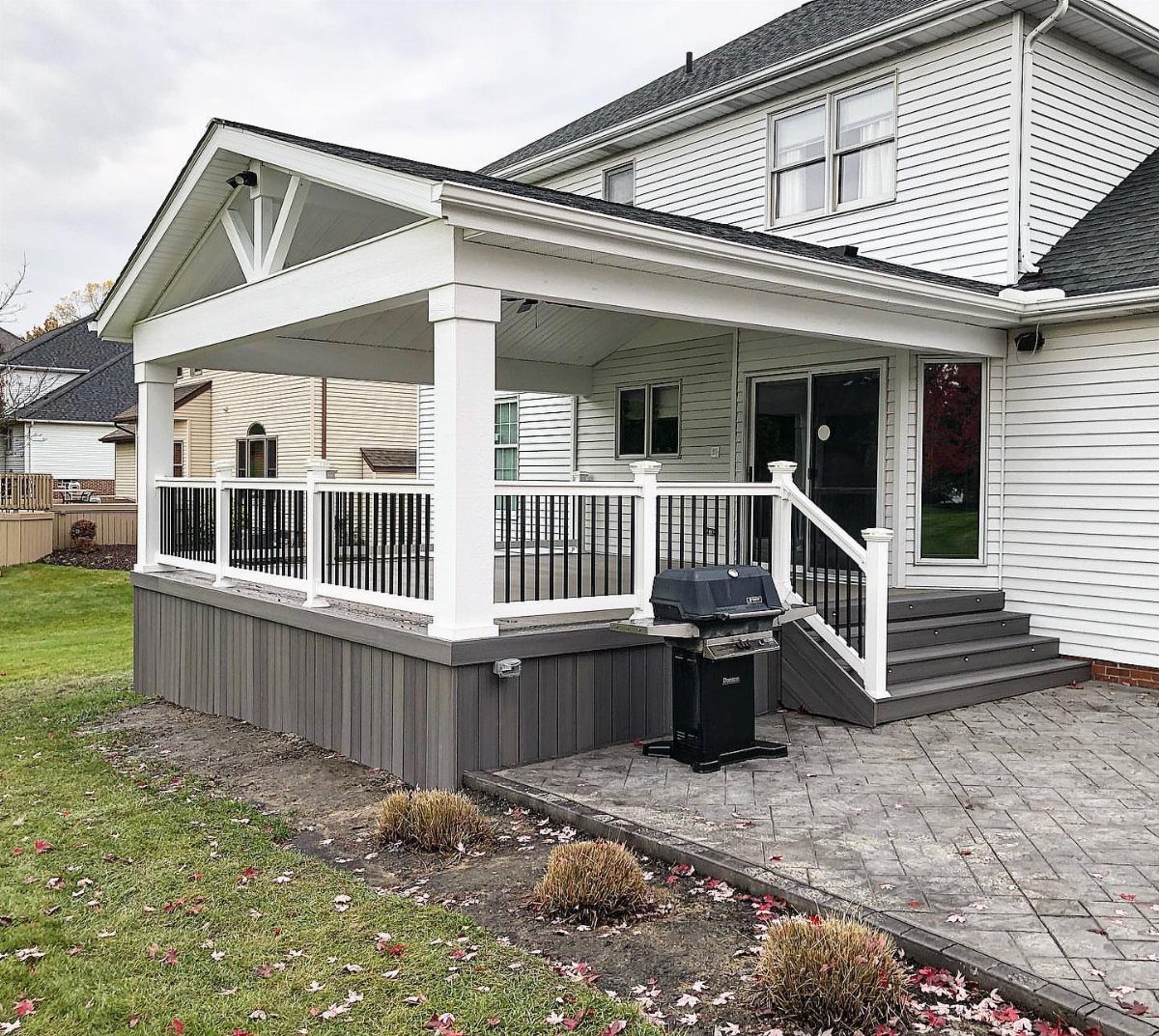 A white covered deck with black railings attached to a white house overlooking a stone patio.
