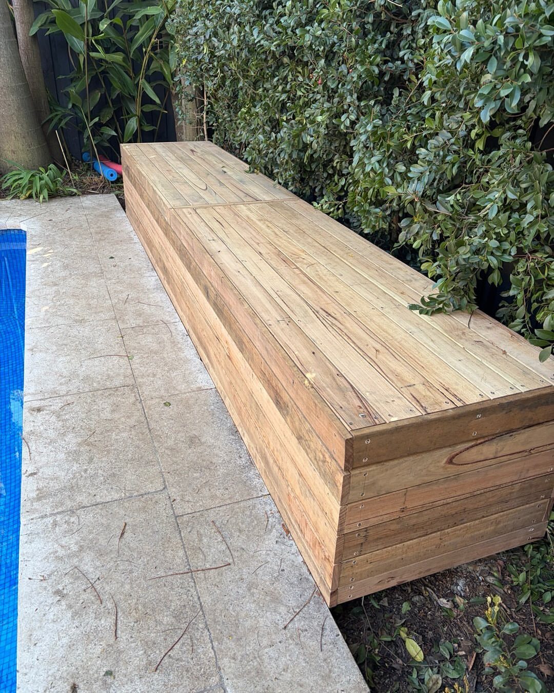 Long wooden storage bench made of light planks sitting on a stone patio outdoors.