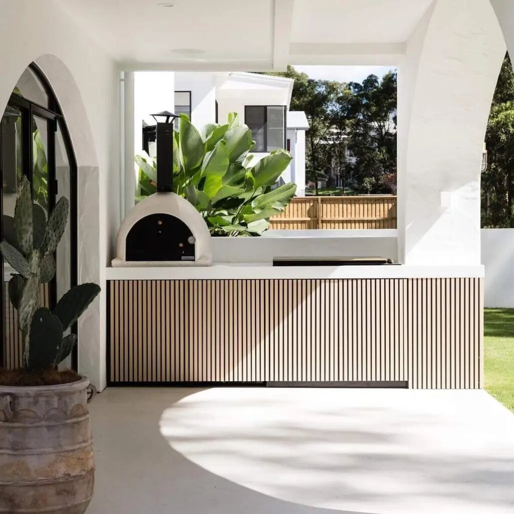 Modern outdoor kitchen with wood slat cabinetry, white countertops, and a dome pizza oven.