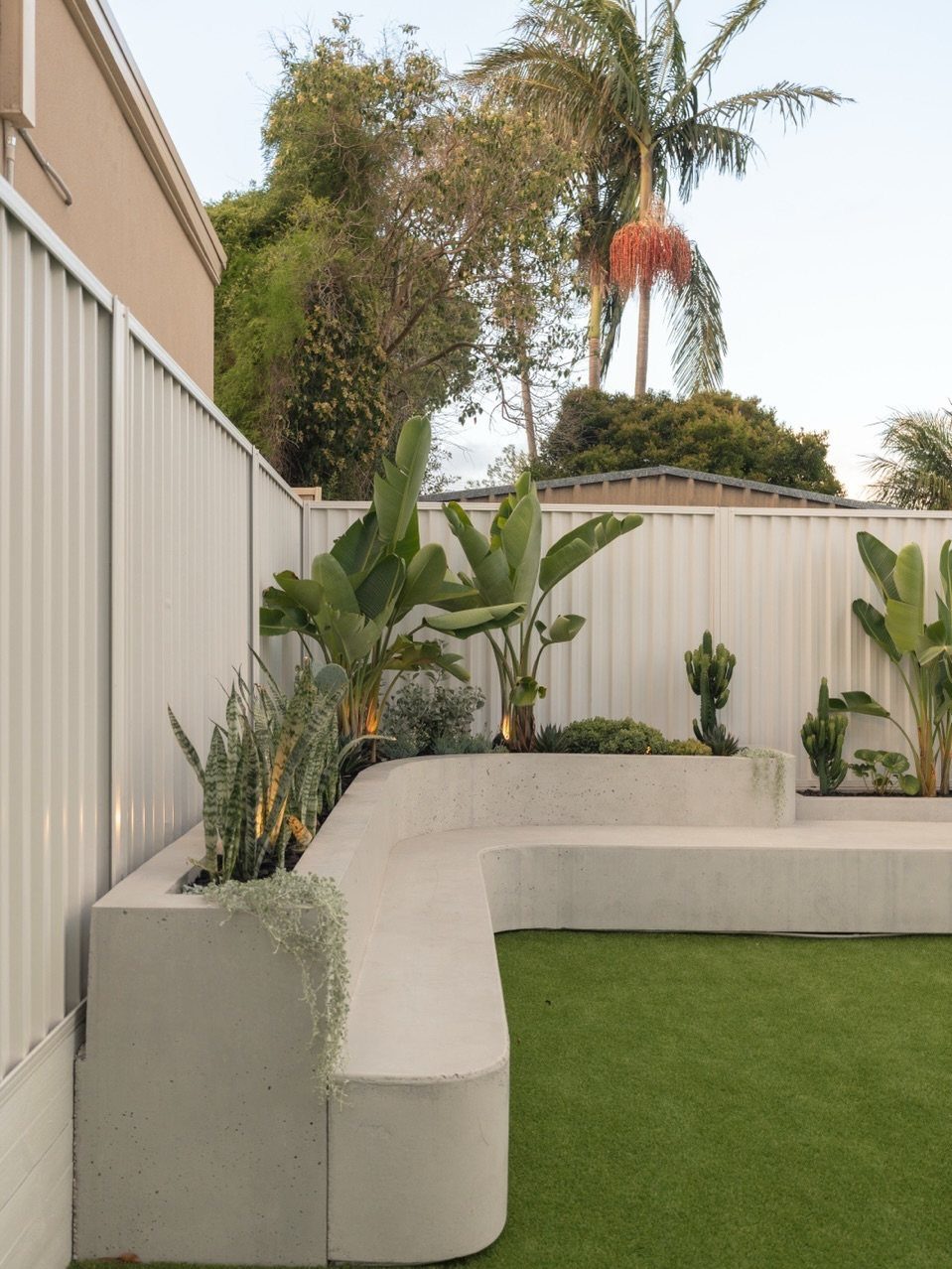 A curved concrete built-in garden bench surrounded by lush tropical plants and green grass.