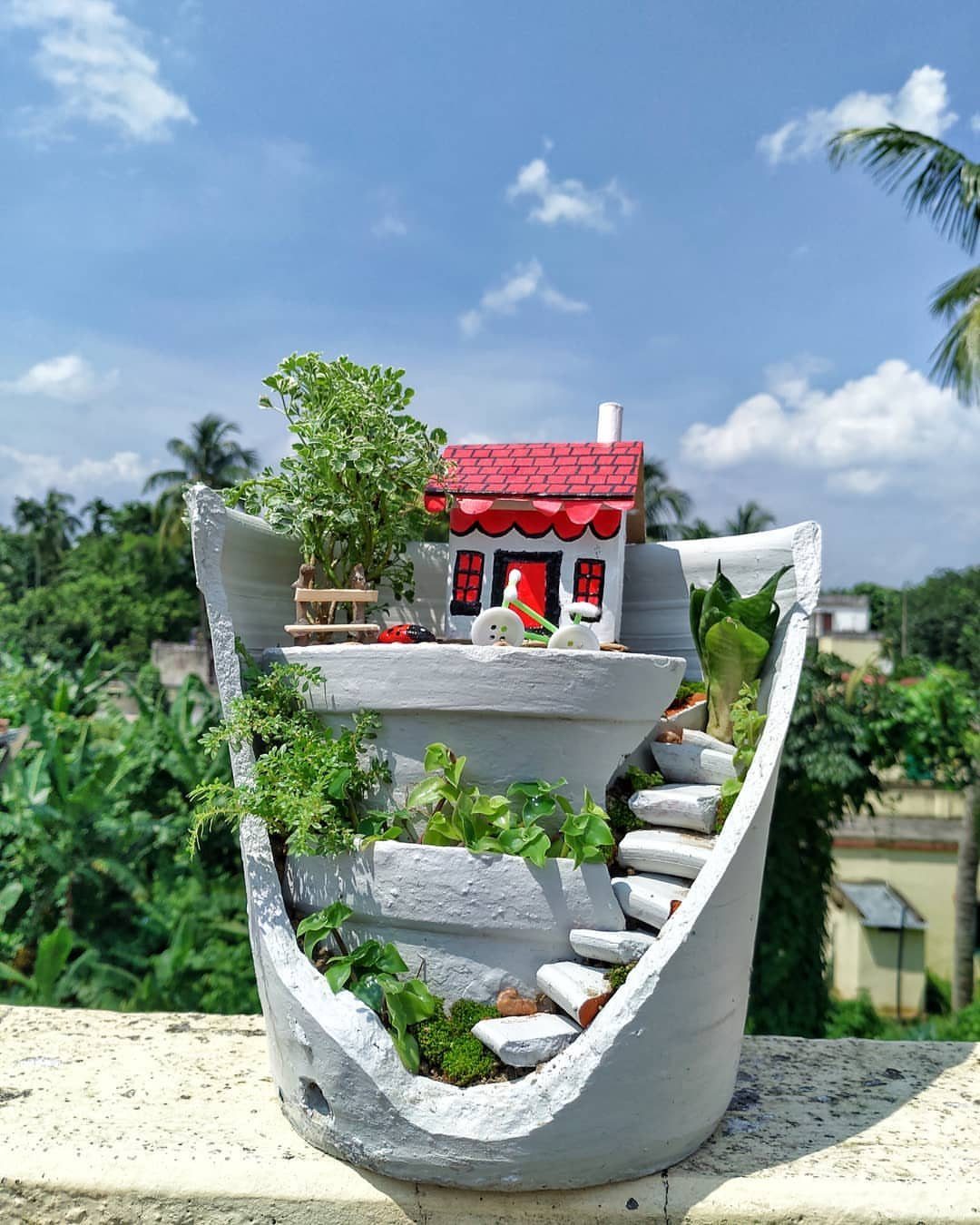 A broken grey pot transformed into a miniature fairy garden with a small house and greenery.