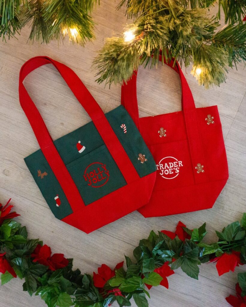 Two small red and green festive fabric tote bags resting on a wooden surface with greenery.