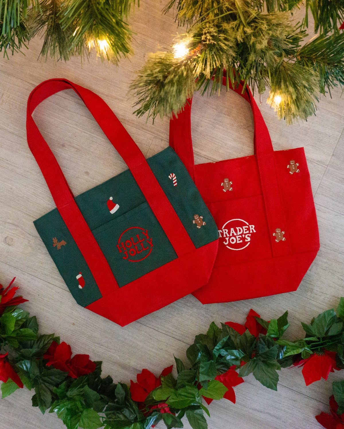 Two small red and green festive fabric tote bags resting on a wooden surface with greenery.