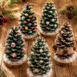 Five decorated pinecone trees with various ornaments sitting on wooden bases on a rustic table.
