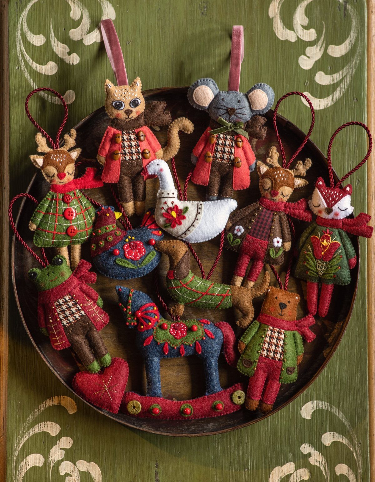 Handmade felt animal Christmas ornaments arranged on a dark wooden tray against a green background.