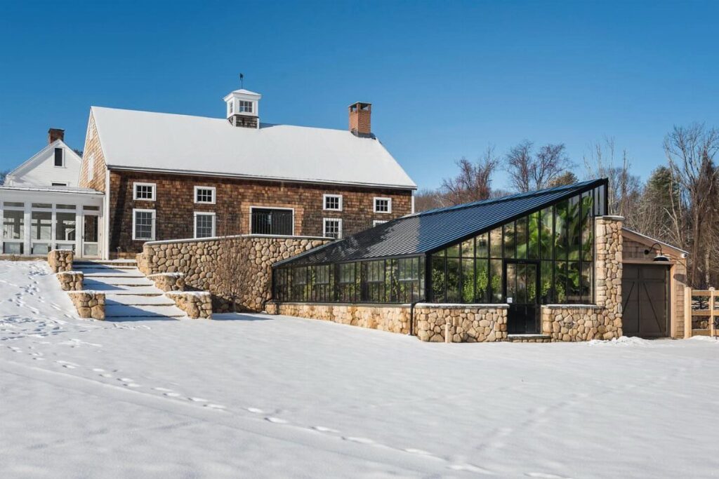A modern greenhouse with a slanted glass roof and stone base sits in a snowy yard.