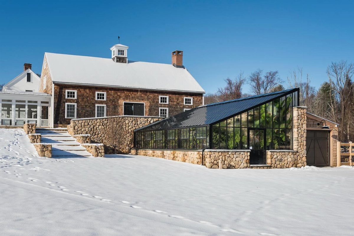 A modern greenhouse with a slanted glass roof and stone base sits in a snowy yard.